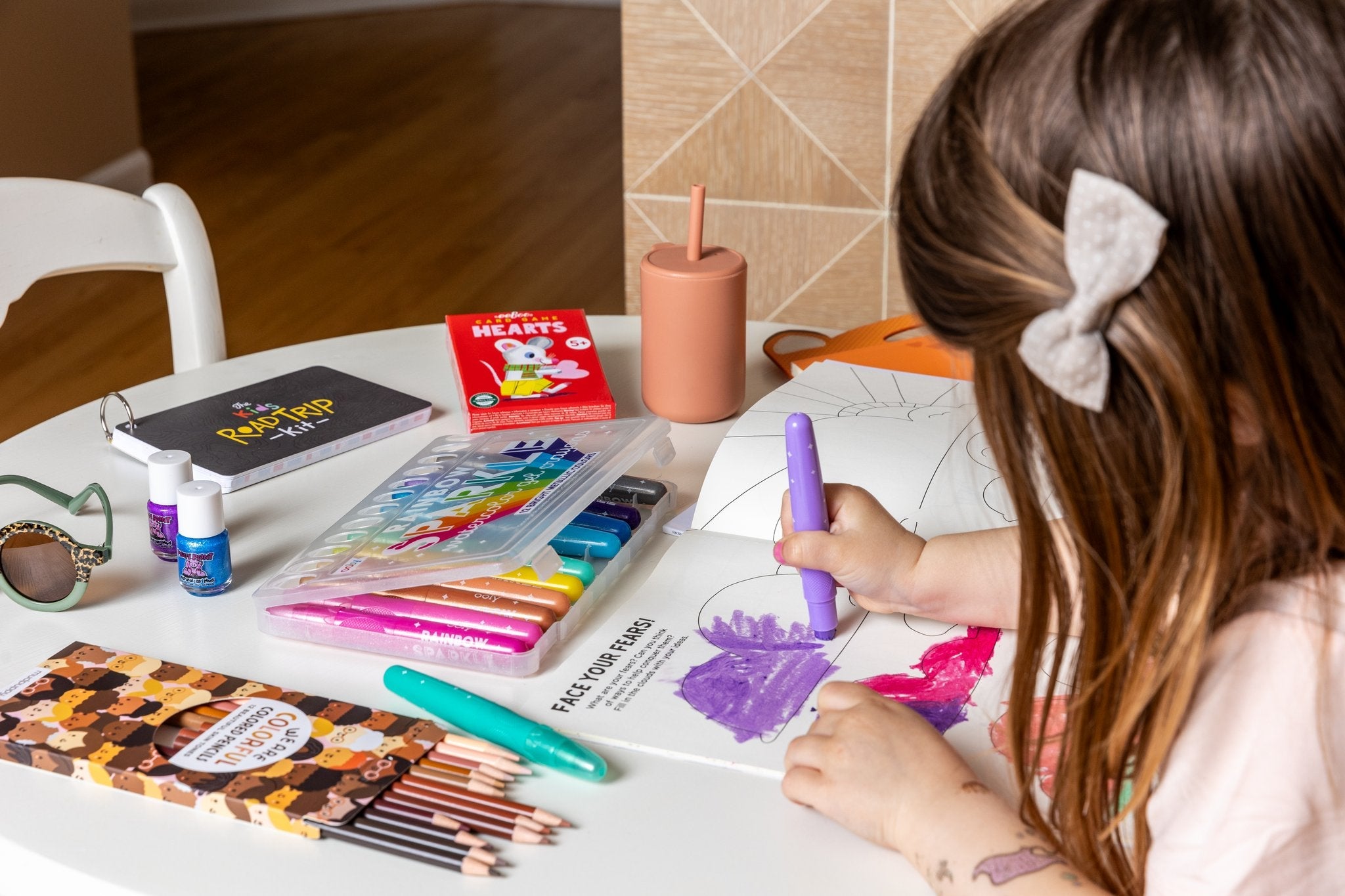 A sweet brunette girl diligently colors in a coloring book on table a strewn with fun kids stuff: a silicone straw cup, nail polish, skin tone colored pencils and fun sunglasses 