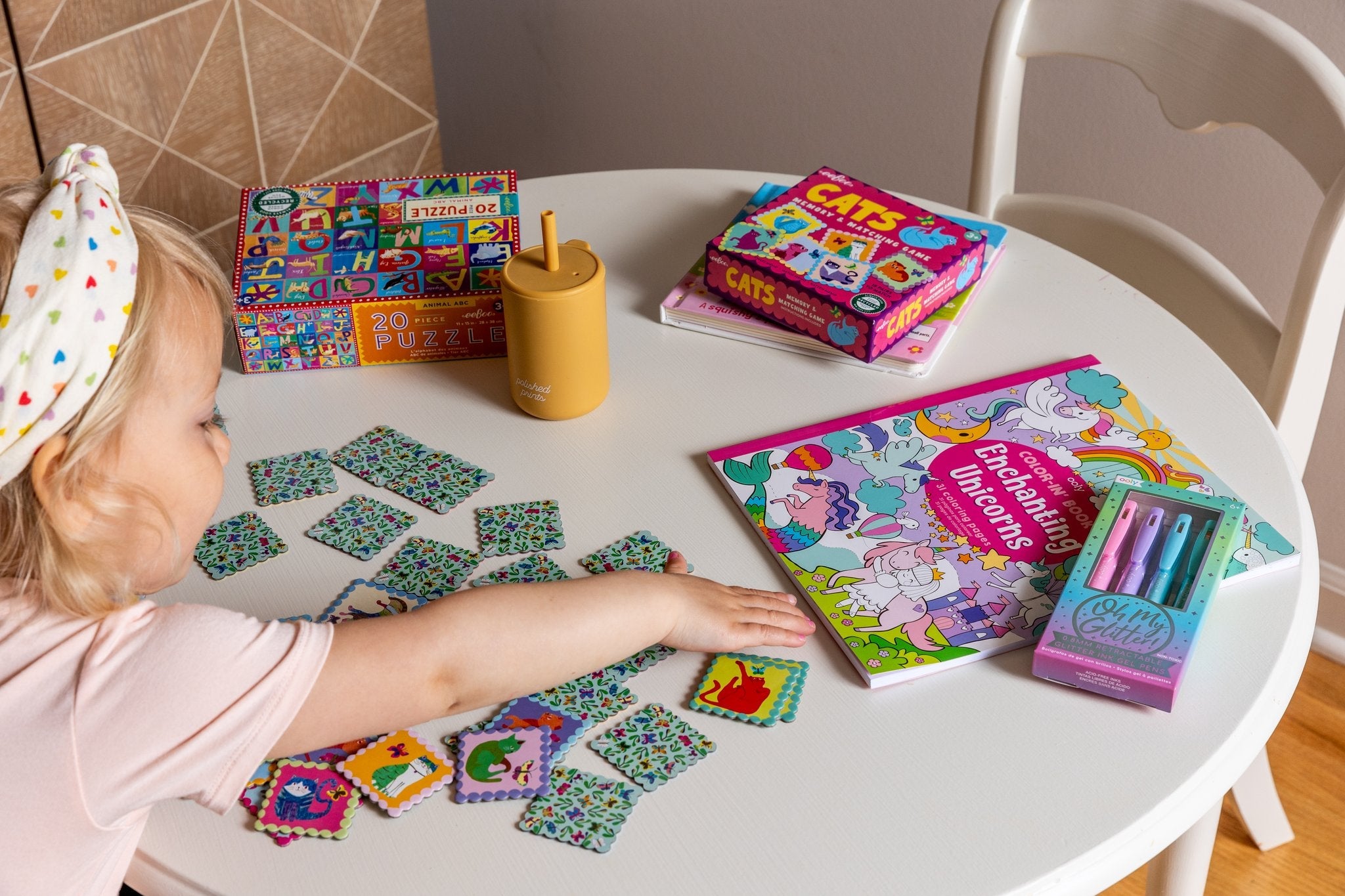 A blonde toddler girl reaches across a table to get a unicorn coloring book across a cat matching game.