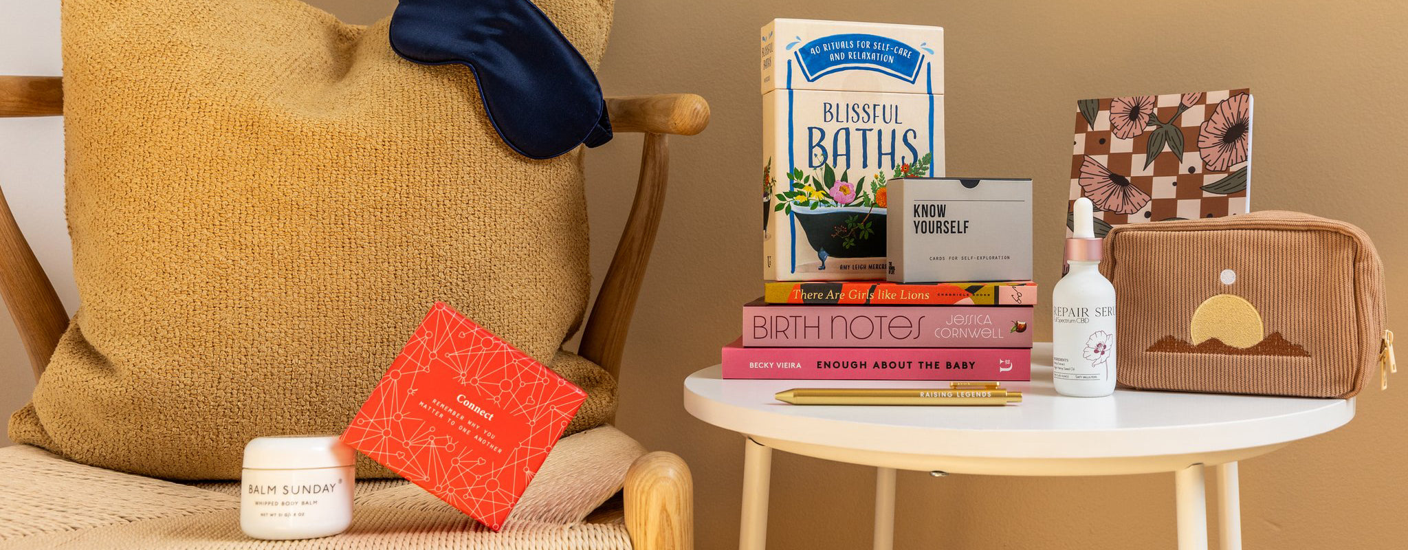 Various gifts—books, serum, a sleep mask, journals, cards, and others—perfect for gifting to a mom, are arranged on a woven chair and white side table.