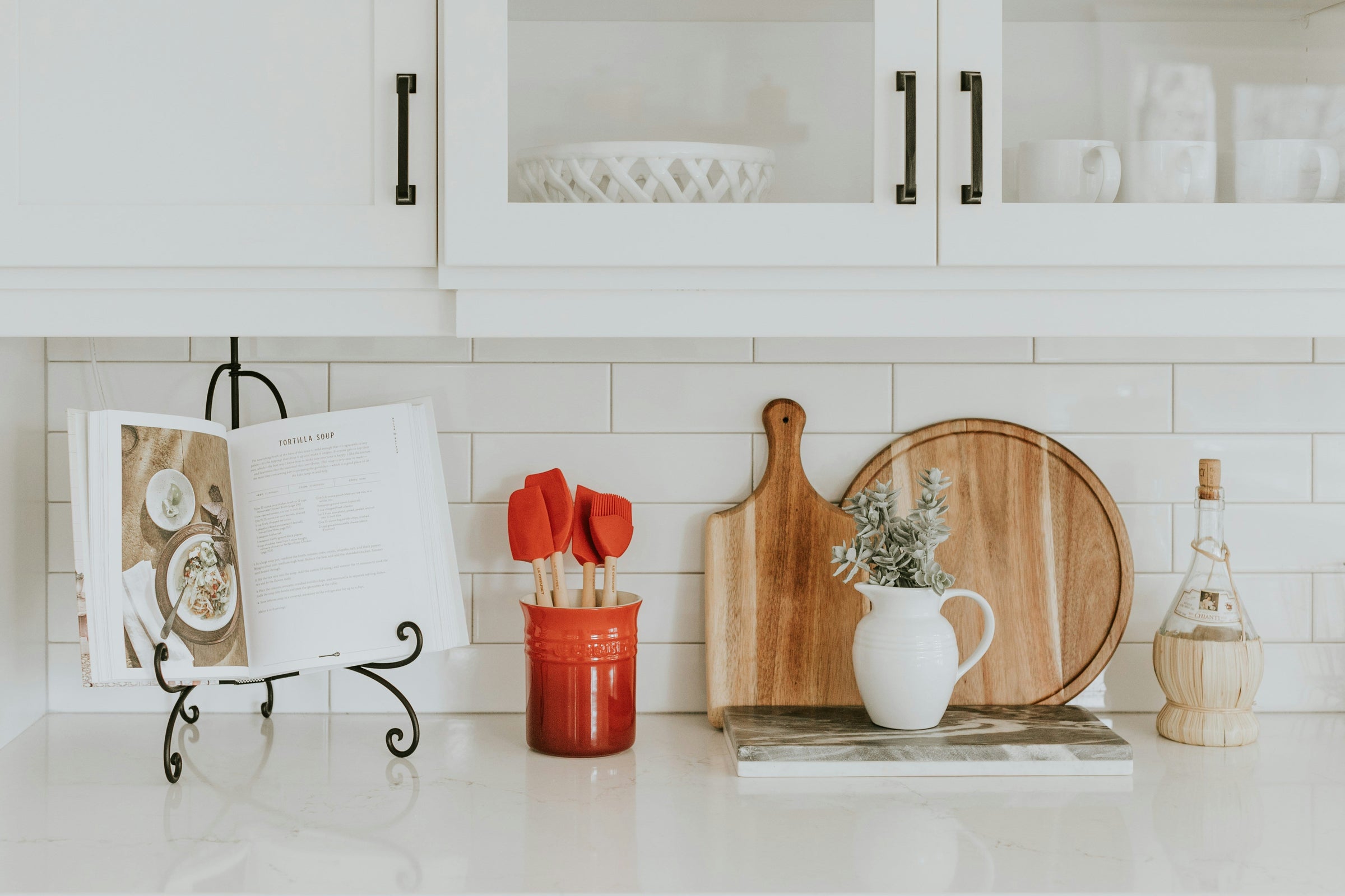 A white kitchen counter is staged with wooden cutting boards, a red crock of kitchen utensils, and an open cookbook on a stand.