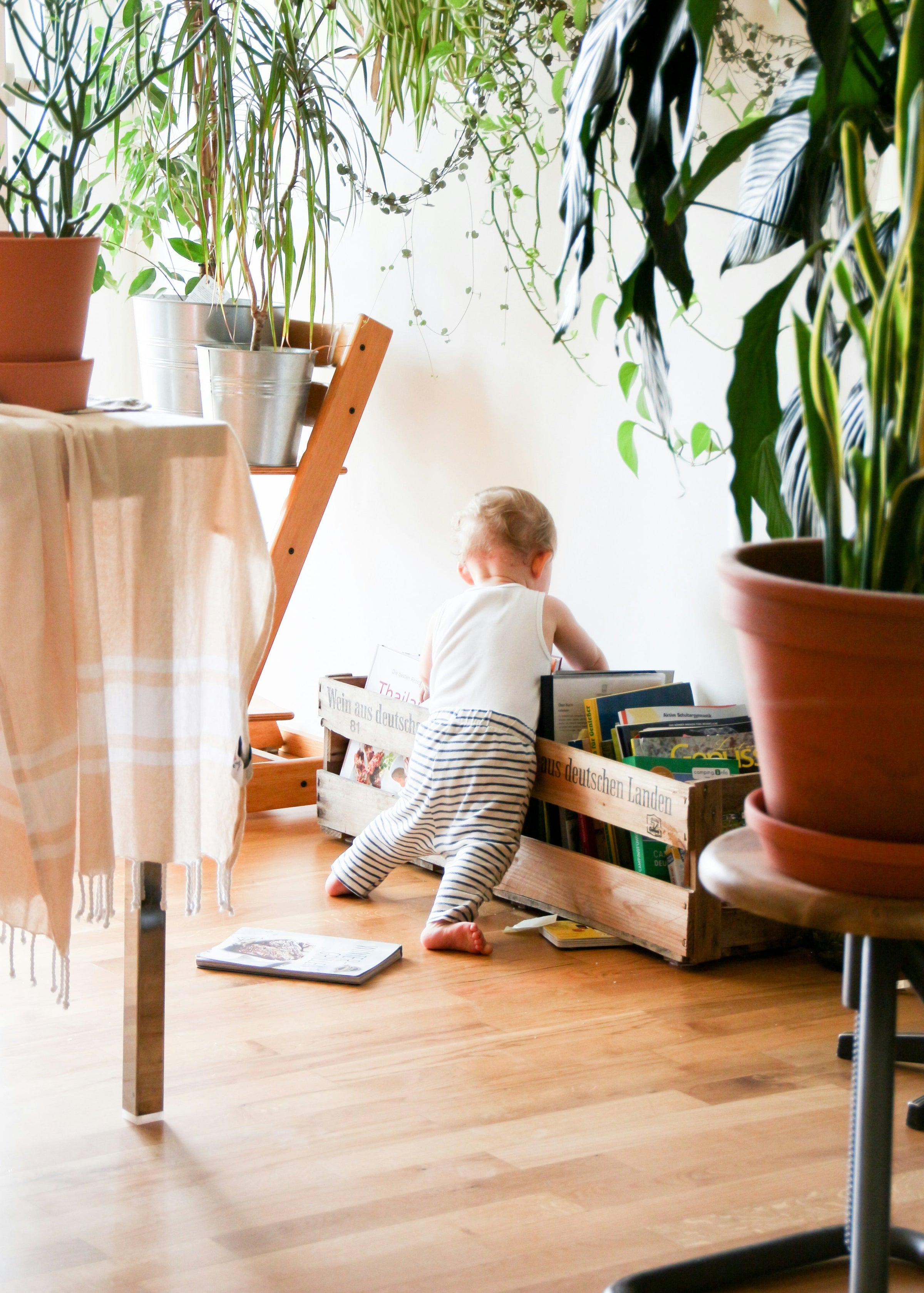 A toddler faces away from the camera, digging in a rustic wooden crate for a book. Other kids books are scattered about amongst simple furniture and houseplants.