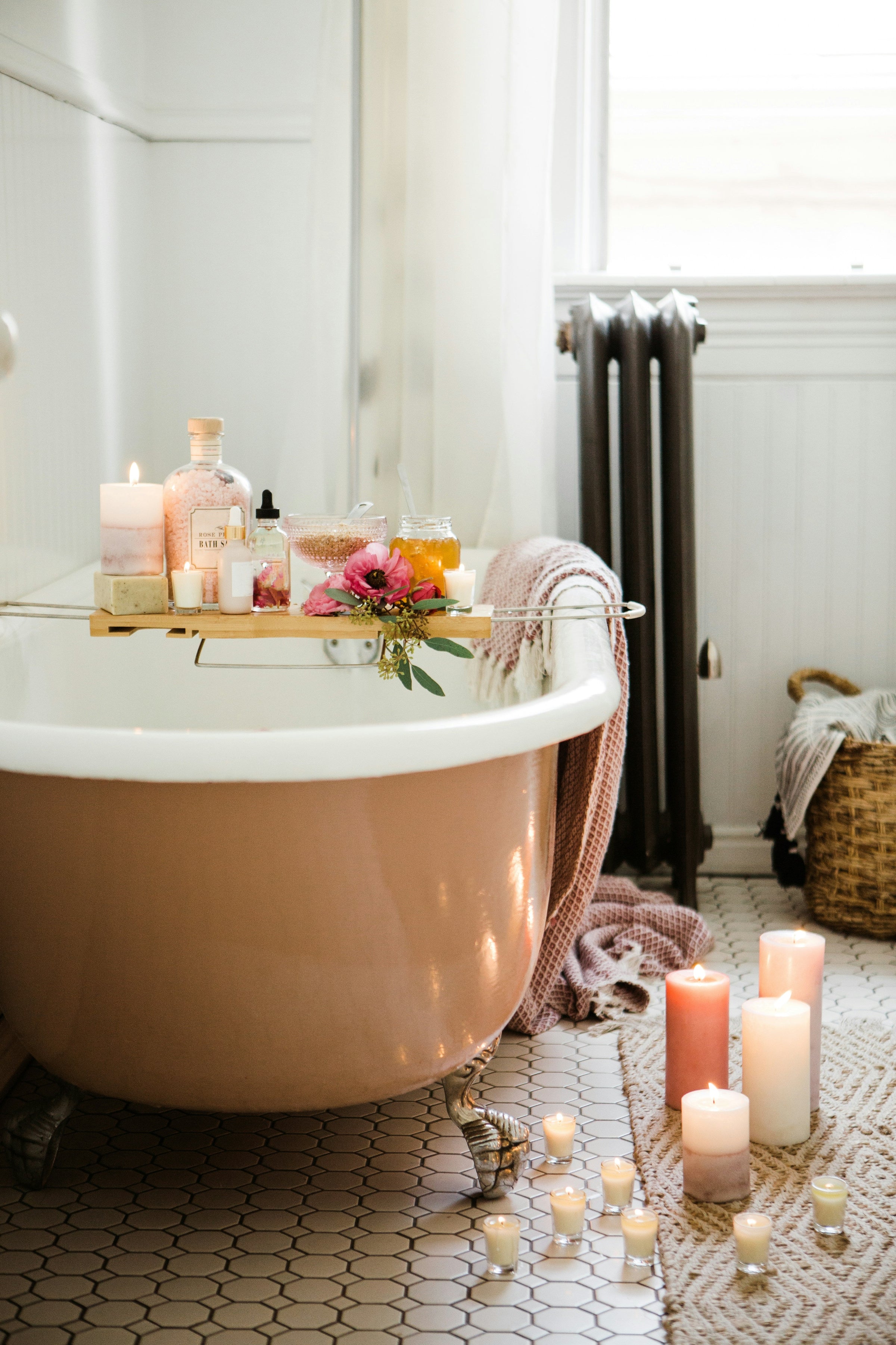 A dusty pink claw foot tub is surrounded by candles. Florals, bath salts, and a soft towel are nearby.