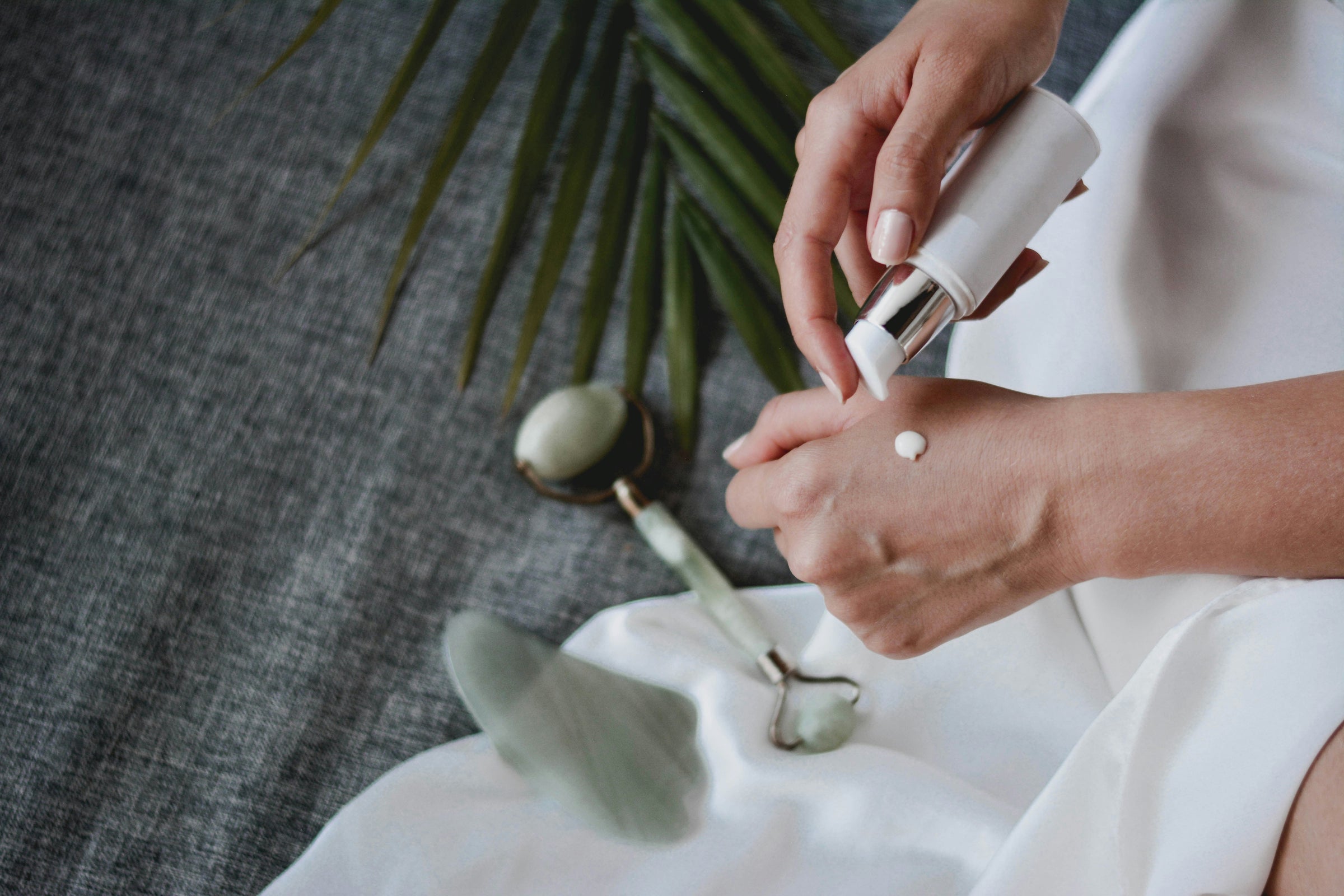 A female hand squirts a small dab of body cream on her hand. A jade roller and palm leaf are below.