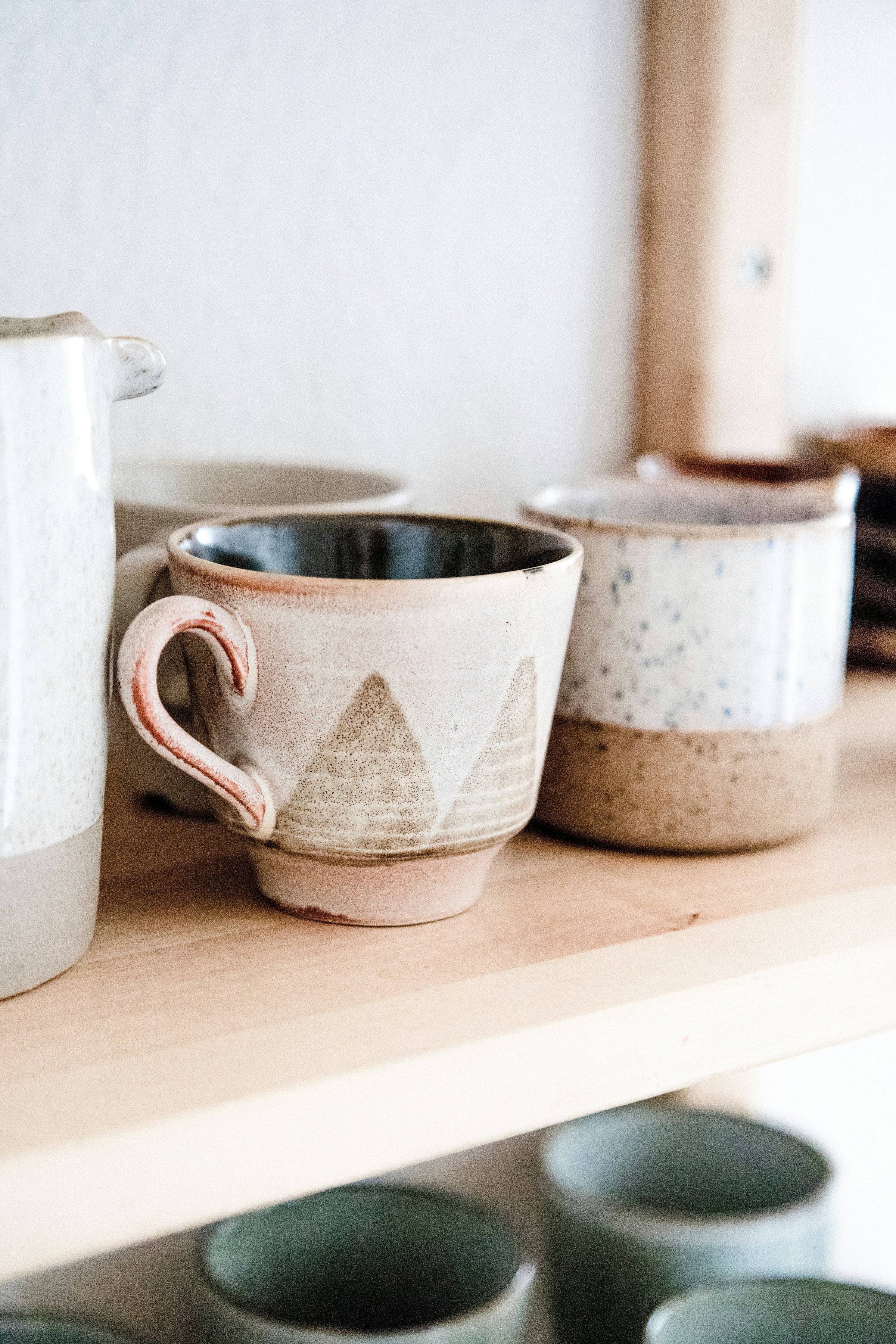 A few handmade white and cream-colored ceramic coffee mugs sit neatly on a wooden shelf.