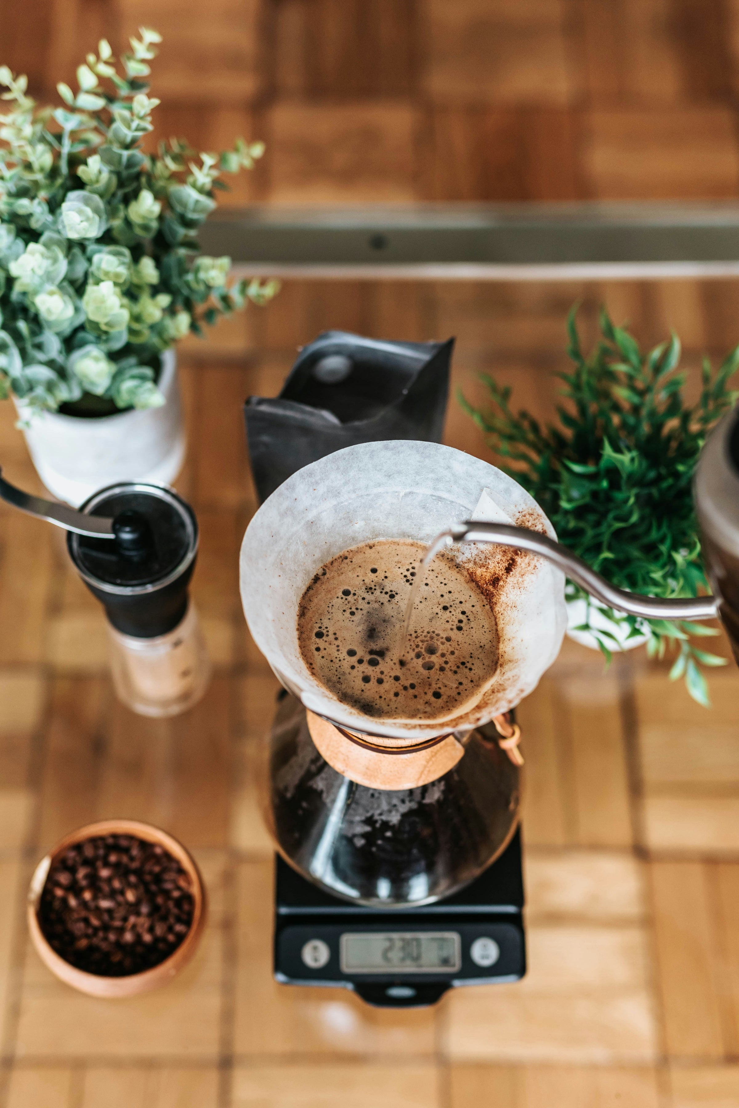 A gooseneck kettle pours hot water over a Chemex coffee maker on top of a gram scale, burr grinder is nearby