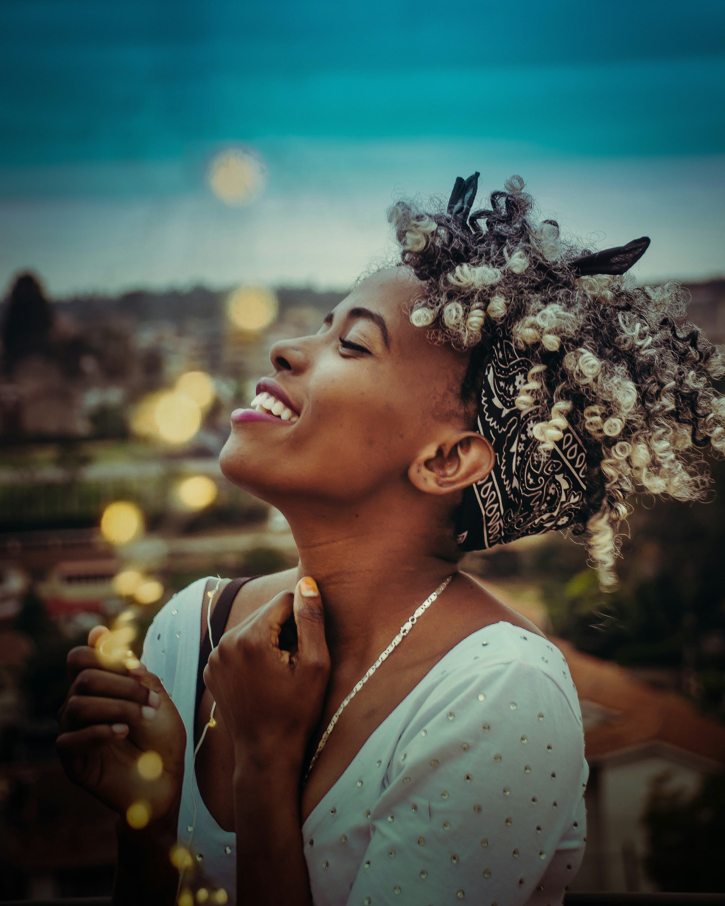 A young black woman with beautiful grey curls tilts her head back towards the sky with joy; her hands near her heart, a smile on her face. A blurry cityscape at twilight is behind her.