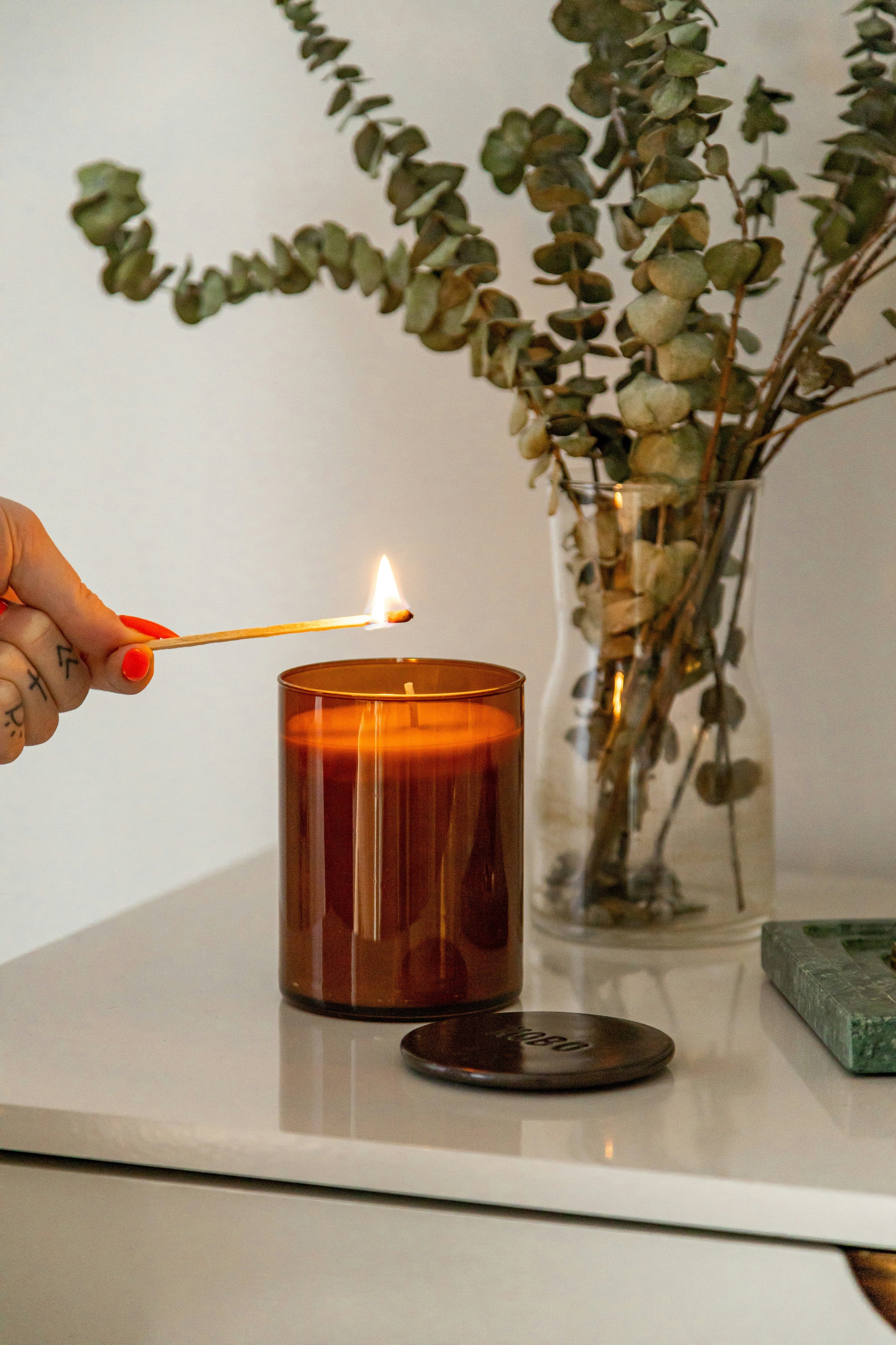 A hand reaches in with a lit match to light an amber glass candle, dried eucalyptus in a vase nearby.