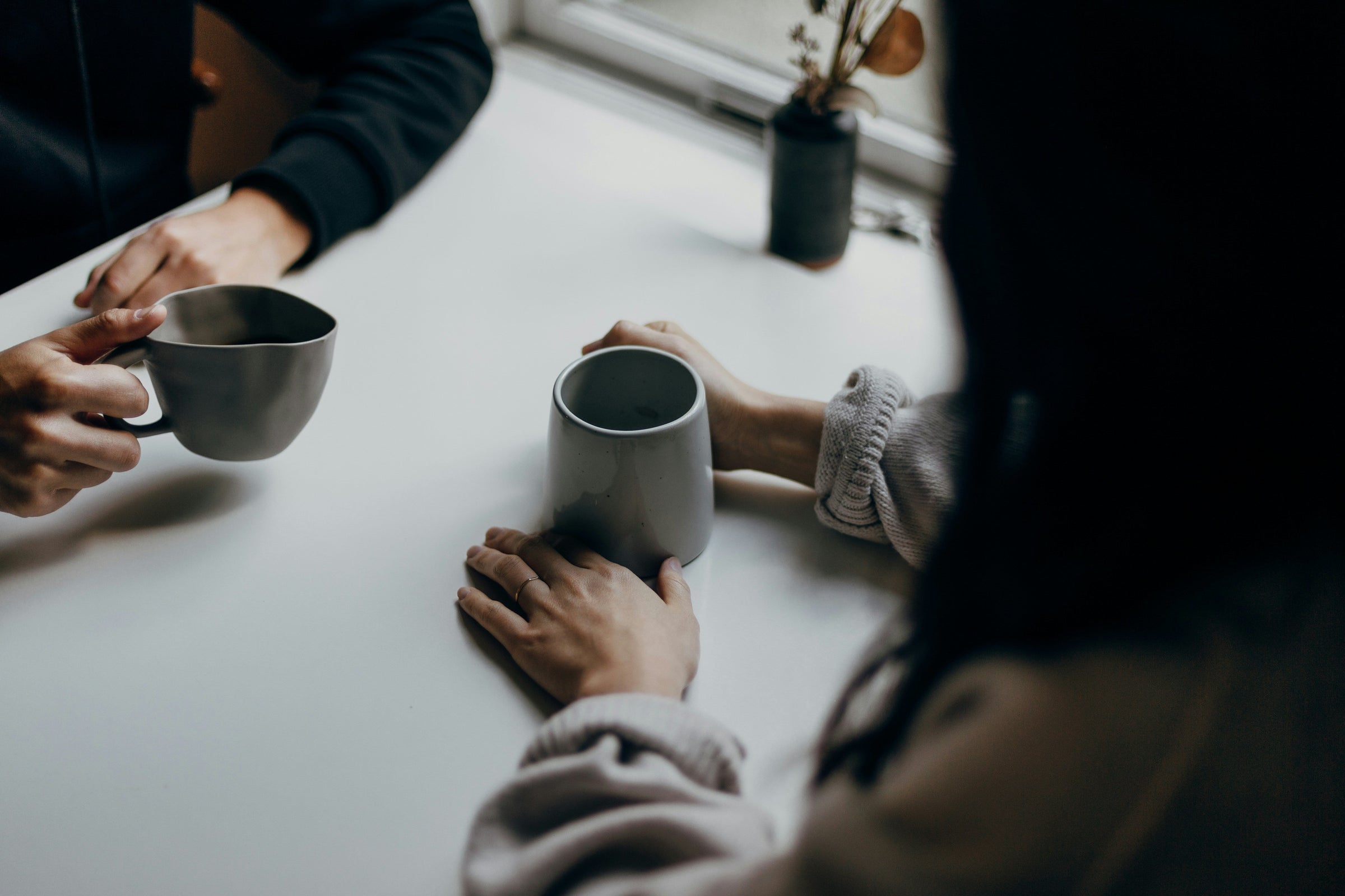 Two people sit across from one another holding differently shaped coffee mugs, the lighting is moody, a simple flower vase is on the table between them.