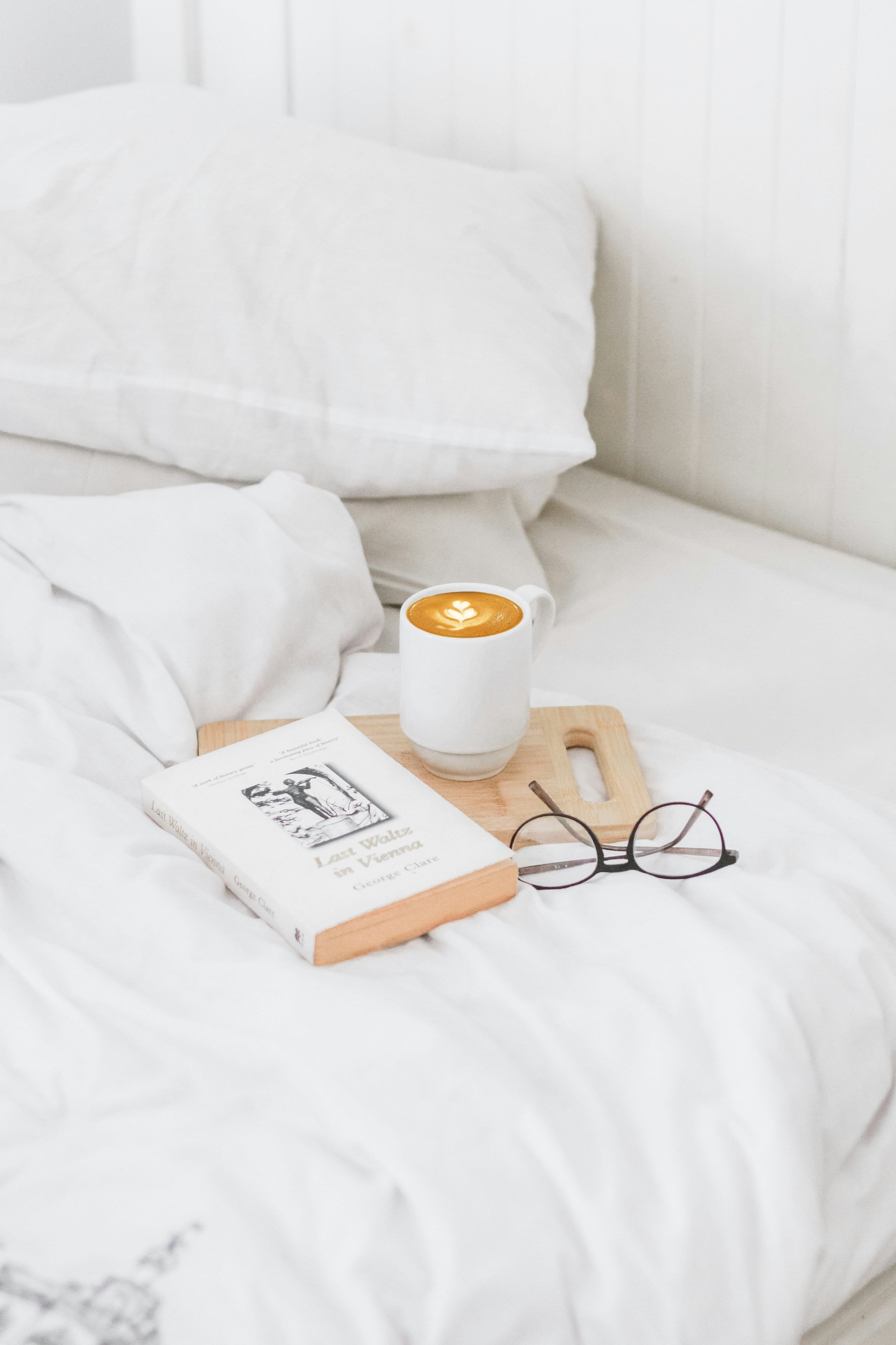 A book, a freshly made latte, and a pair of reading glasses are arranged on a white duvet cover near a fluffy white pillow.