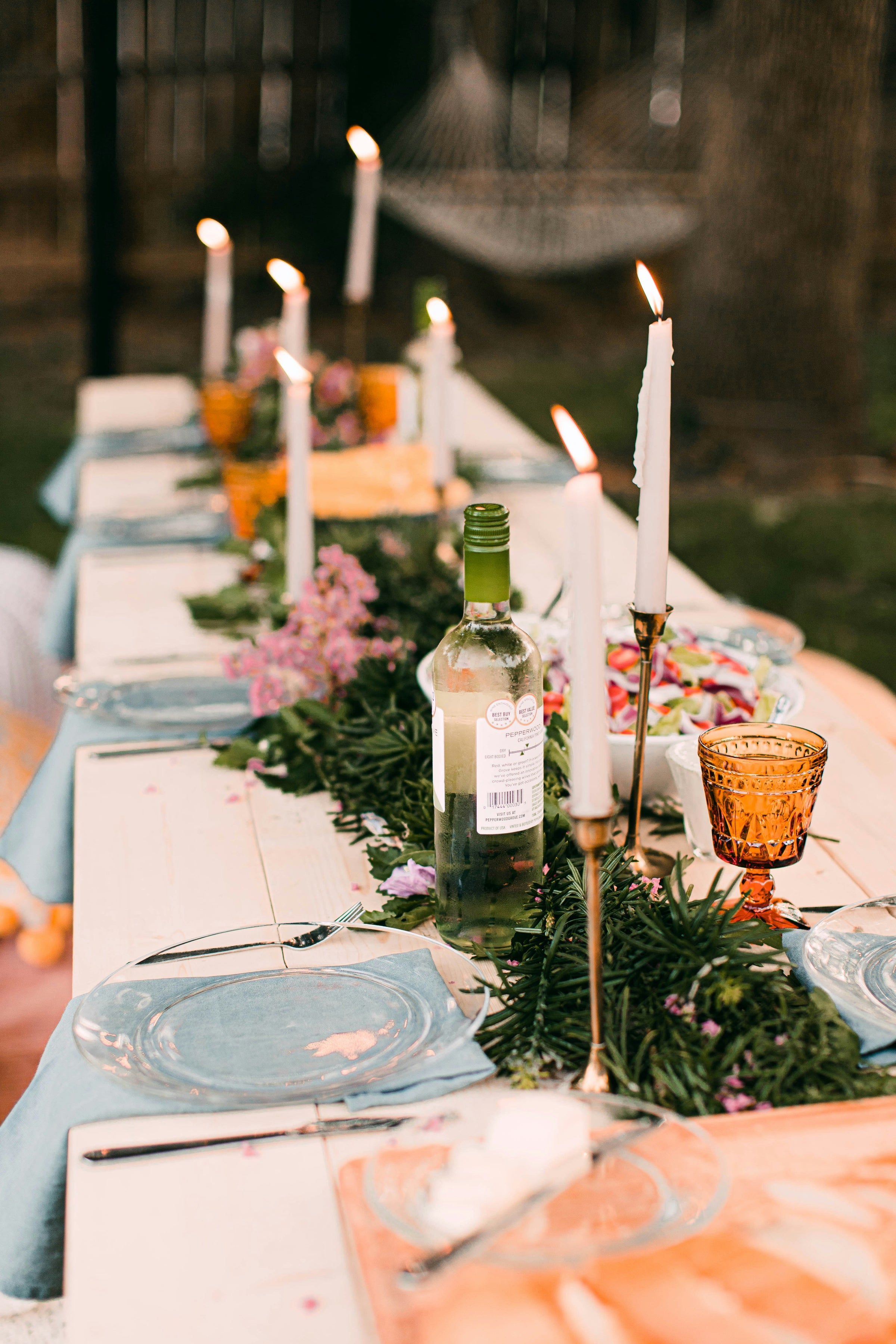 A stunning long table is nicely dressed with long taper candles, clear glass plates, fresh greenery, vintage glasses, perfectly set for a dinner party.