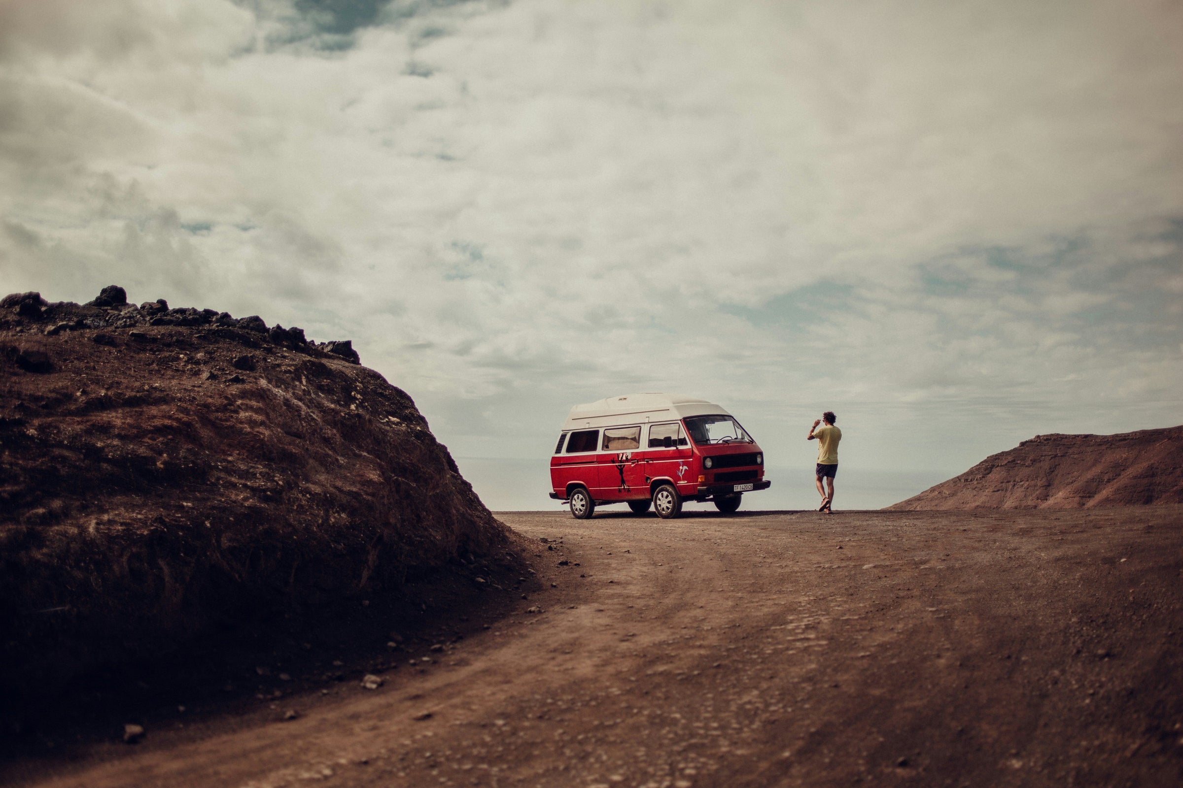 On a mountaintop lookout stop, a person stands near a old red travel van to take a photo of the stunning view.