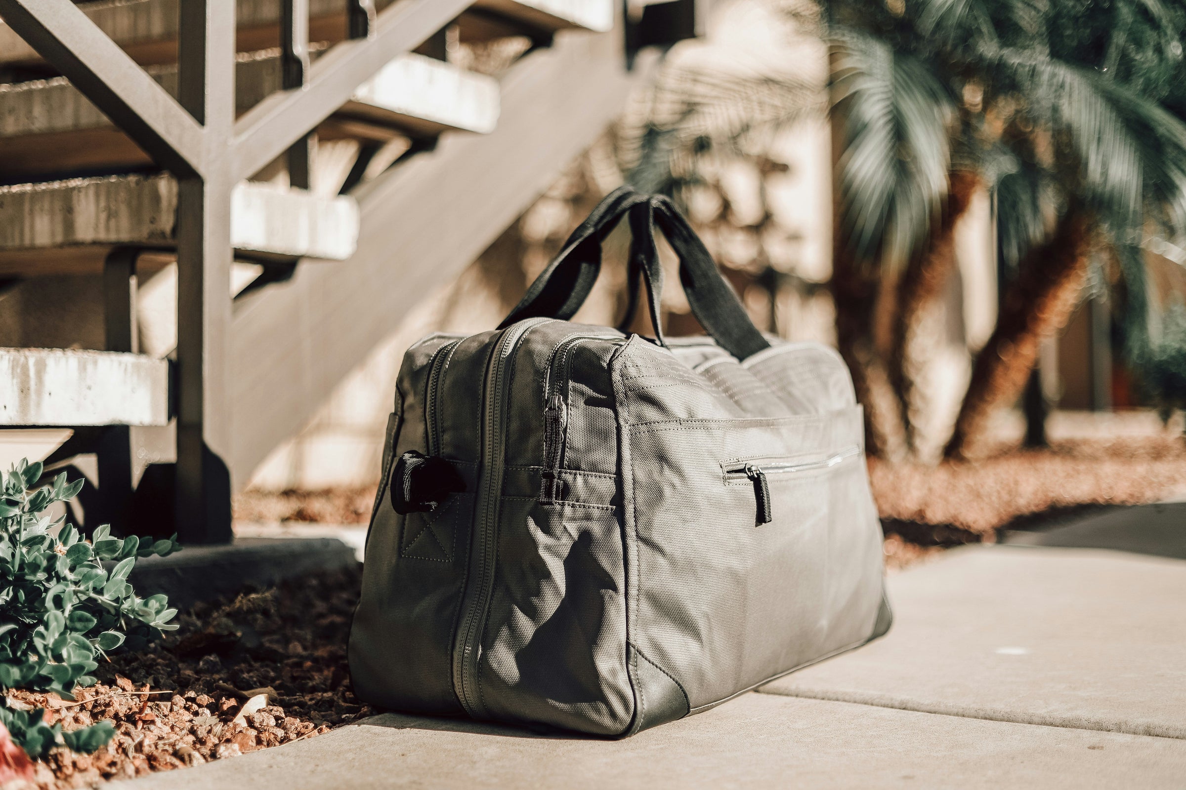 A cool, modern grey-green duffle bag sits outside on a sidewalk near stairs.